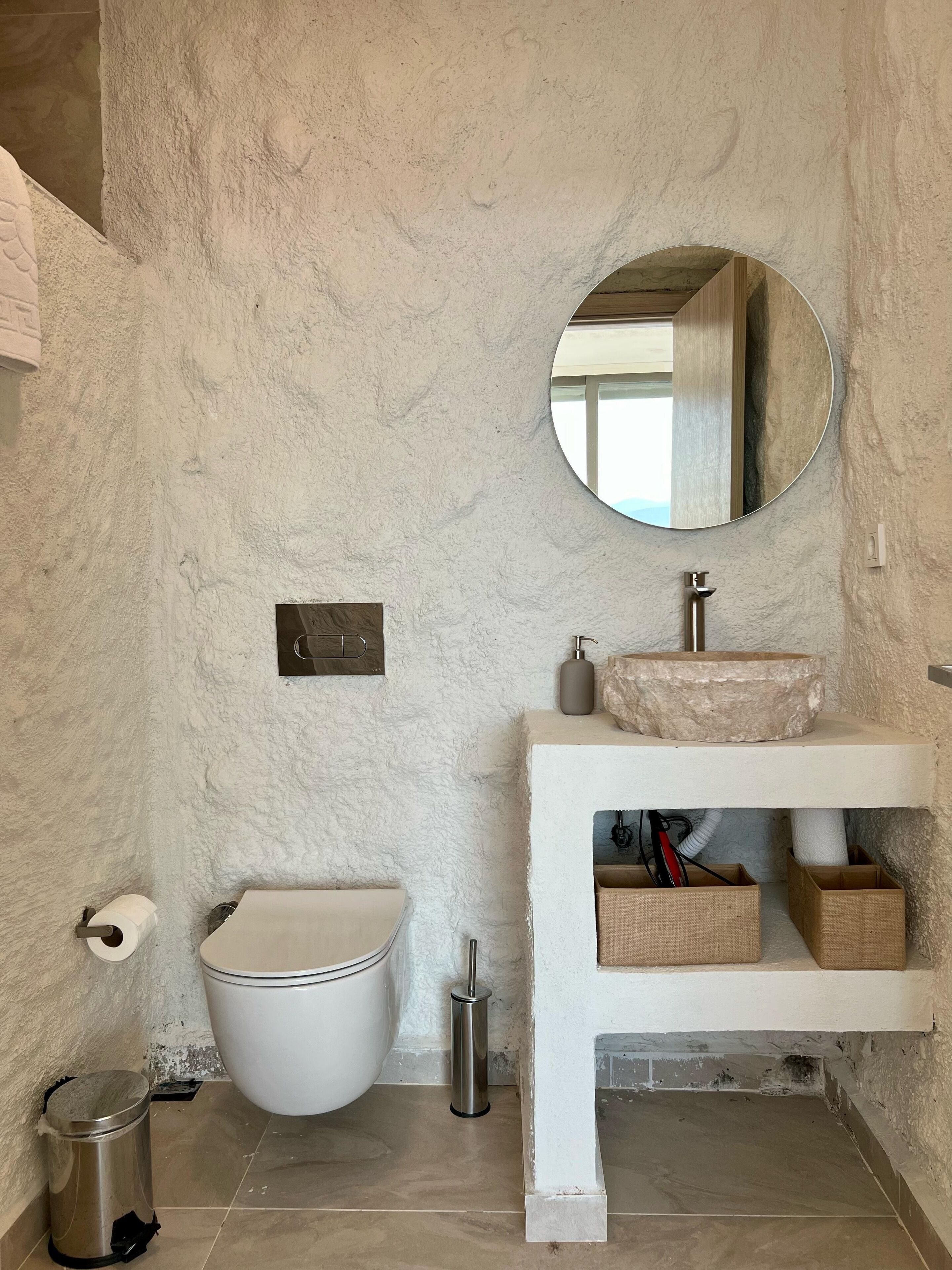 A bathroom with a white textured wall, a round mirror, a white sink with a stone basin, a toilet, and a trash bin.