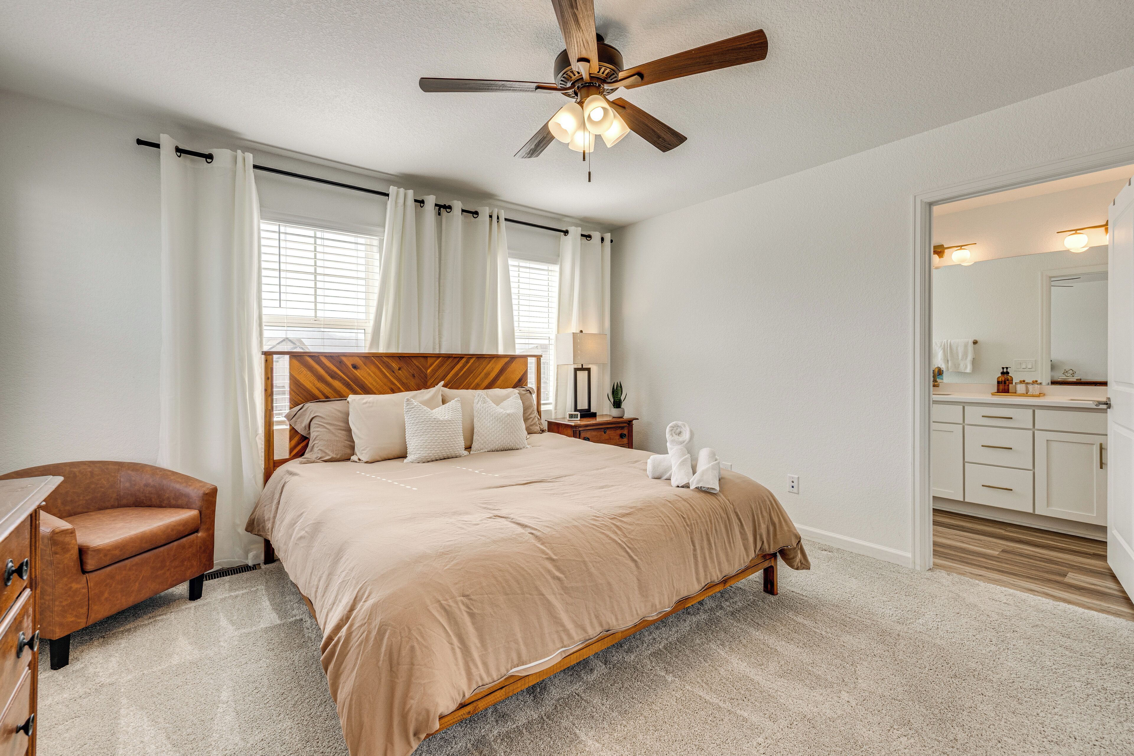 A bedroom with a bed, a chair, a nightstand, a ceiling fan, and a bathroom visible through an open door.