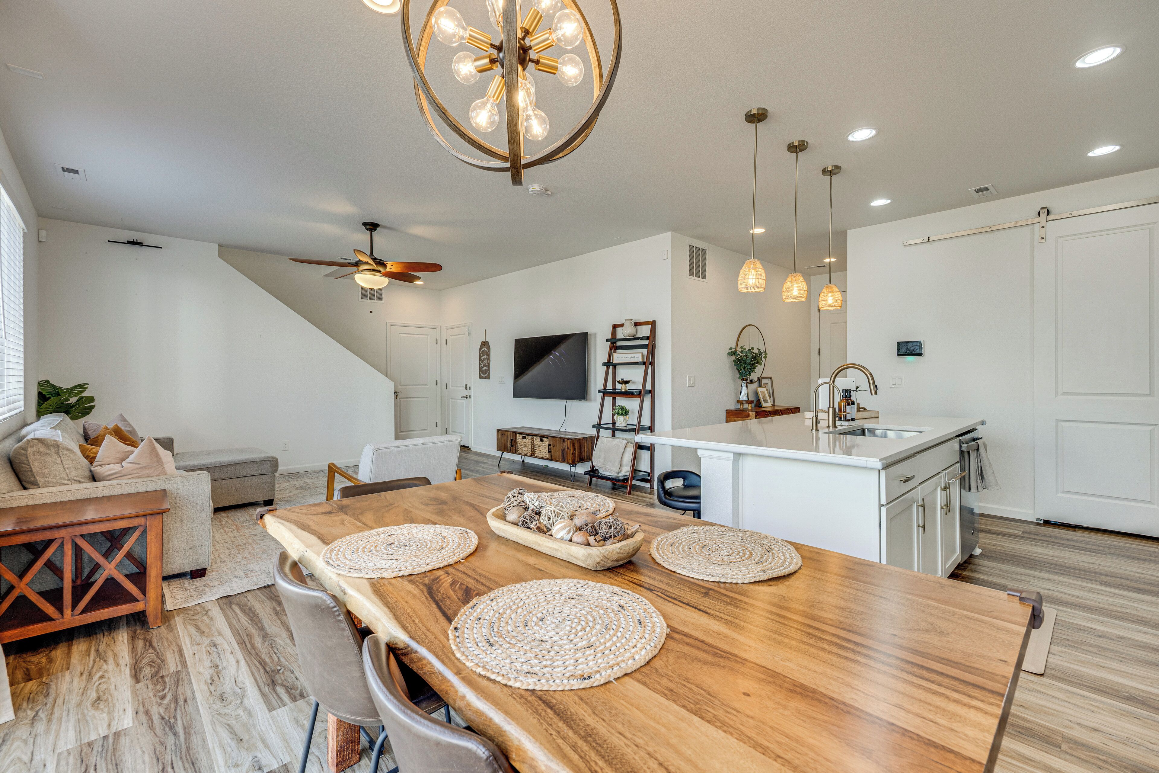 A modern kitchen with a central island, a dining area with a wooden table, and a living room with a sofa and a ceiling fan.