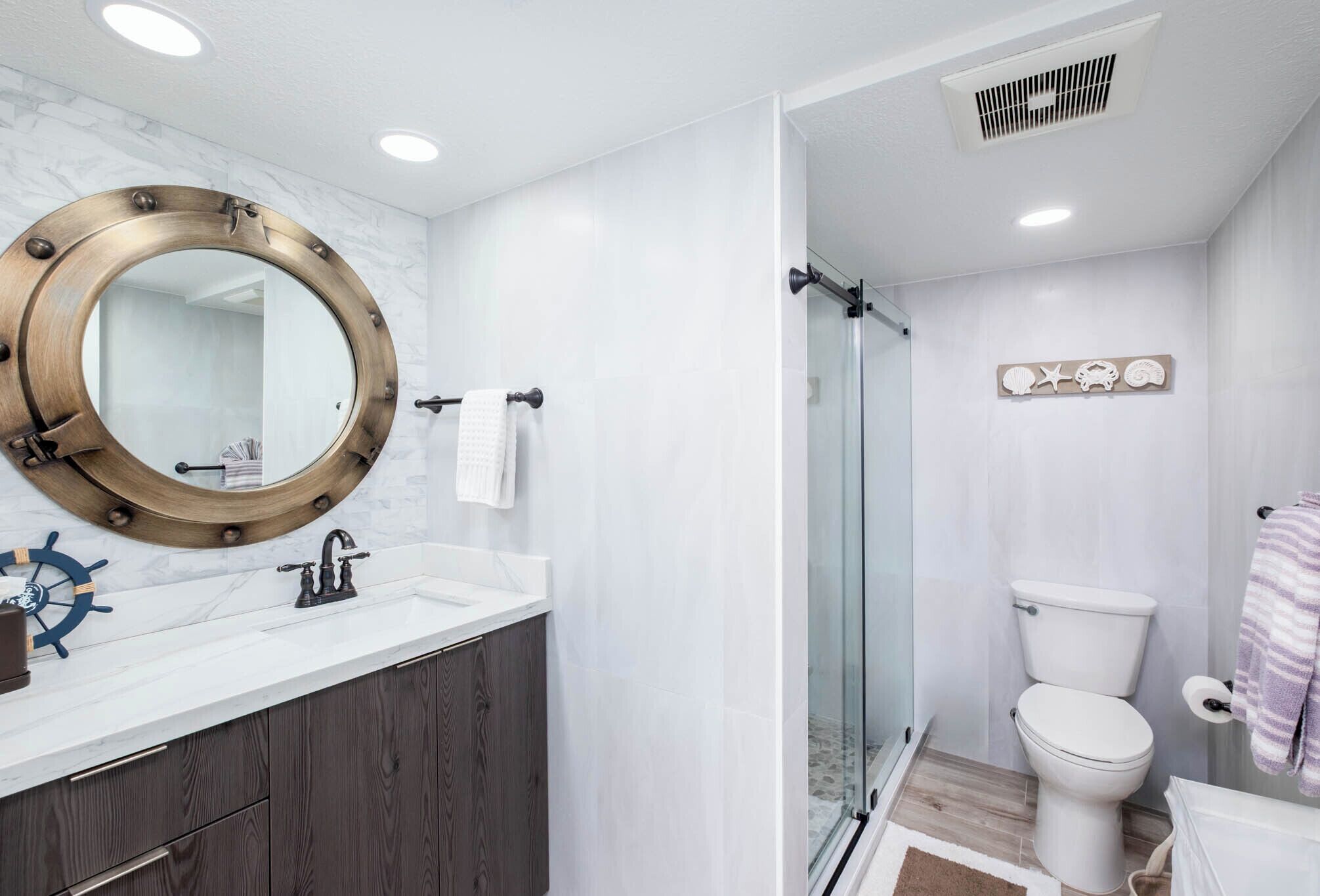 A bathroom with a round mirror, a ship's wheel decoration, a modern sink, and a glass-enclosed shower.