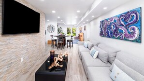 A modern living room with a flat-screen TV, a sofa, a coffee table with decorative items, and a dining area with chairs and a table. - Mariners Club Unit 904, VRNR Luxury Collection, Brand New Completely Remodeled (Key Largo)