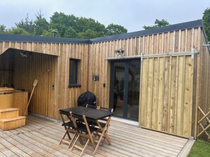 A wooden cabin with a deck, a table and chairs, a grill, and a sliding glass door. - Côté jardin Cottage (near Dinan) (Bobital)