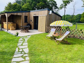 A wooden cabin with a patio area, a hot tub, and a seating area with a striped umbrella. - Côté jardin Cottage (near Dinan) (Bobital)