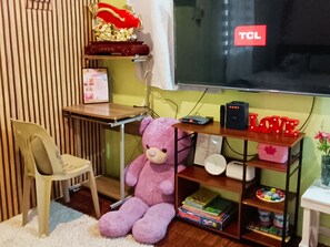 A room with a desk, a chair, a large teddy bear, a shelf with books and a clock, and a TCL TV. - Amigas Transient House (Santo Tomas)