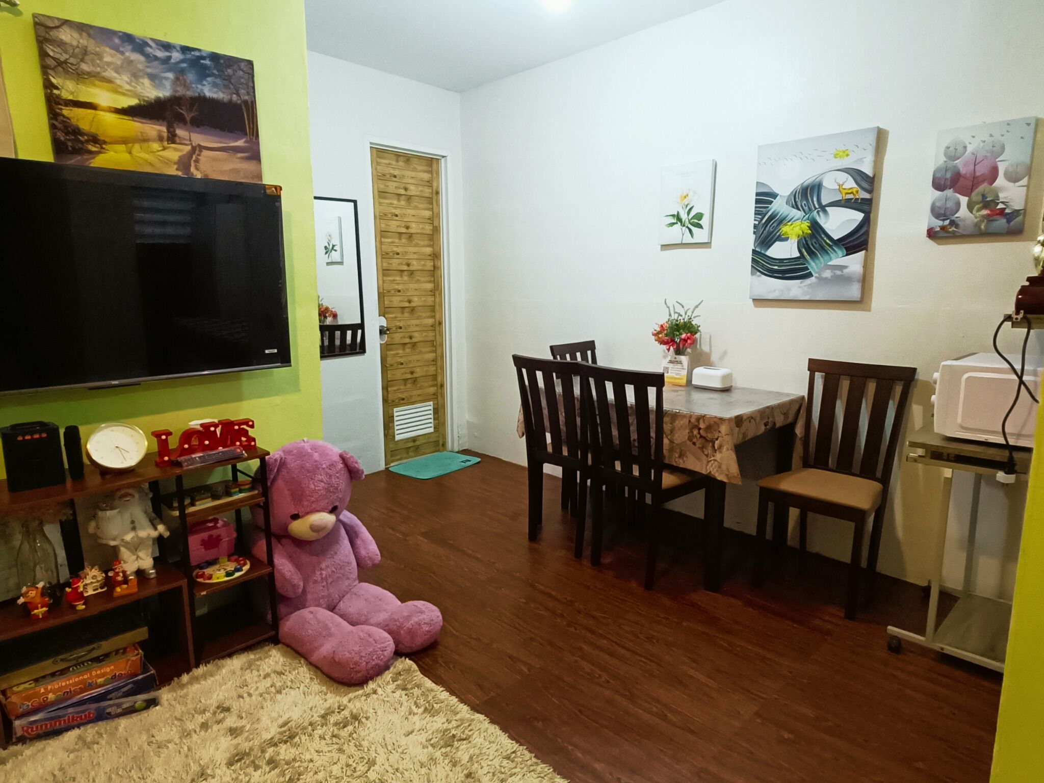 Una sala de estar con televisor, un gran oso de peluche, una mesa de comedor con sillas y obras de arte en las paredes.