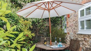 A wooden table with a white umbrella, surrounded by chairs with green cushions, set against a stone wall with greenery.
