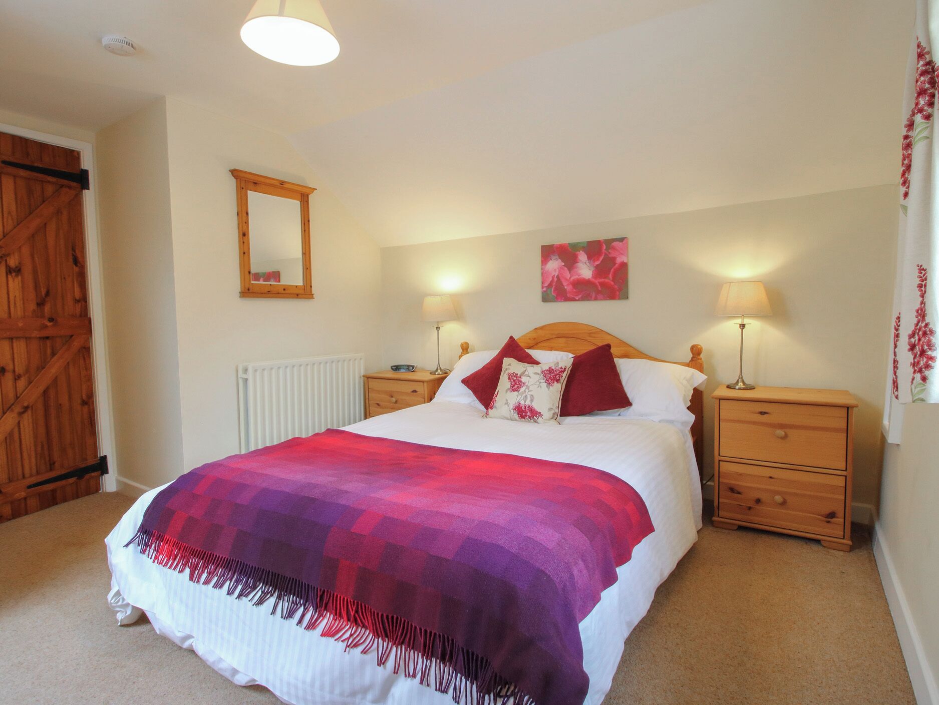 A bedroom with a bed, two bedside tables, a wooden wardrobe, a mirror, and a floral artwork on the wall.