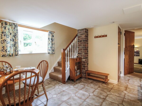 A dining area with a wooden table and chairs, a staircase with a brick wall, and a view of the outdoors through a window with floral curtains. - Little Barn (Oswestry)