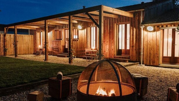 Een rustiek houten gebouw met een vuurkorf op de voorgrond, omringd door zitplaatsen en grind.