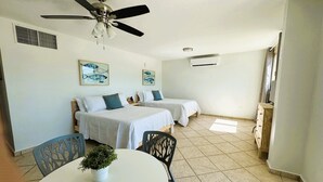 Une chambre avec deux lits, un ventilateur de plafond, une petite table avec une plante et un téléviseur posé sur une commode en bois.