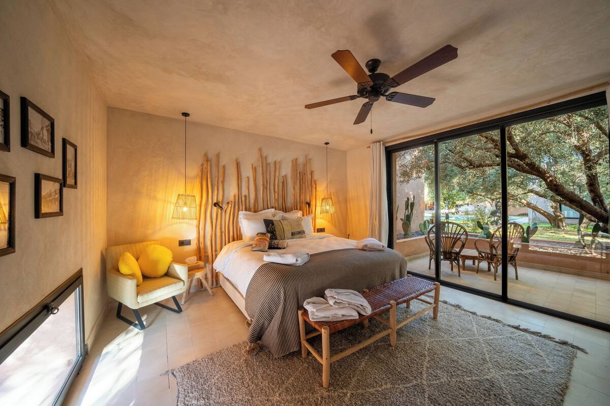 A modern bedroom with a large bed, a ceiling fan, and a view of the outdoors through large windows.