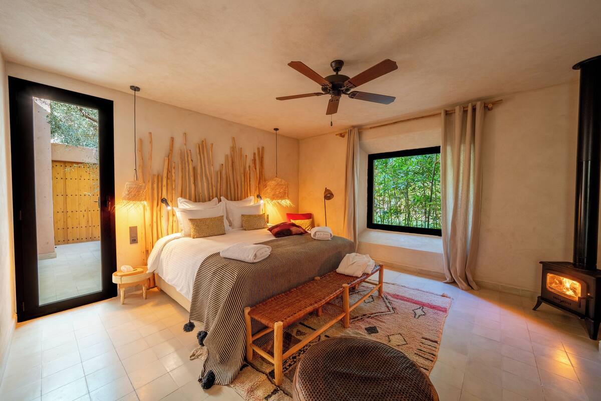 A bedroom with a bed, bedside lamps, a bench, a window with a view, a ceiling fan, and a wood-burning stove.