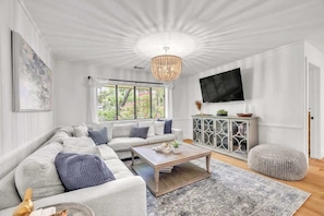 A living room with a sectional sofa, a coffee table, a TV mounted on the wall, a cabinet with decorative items, and a window with blinds.
