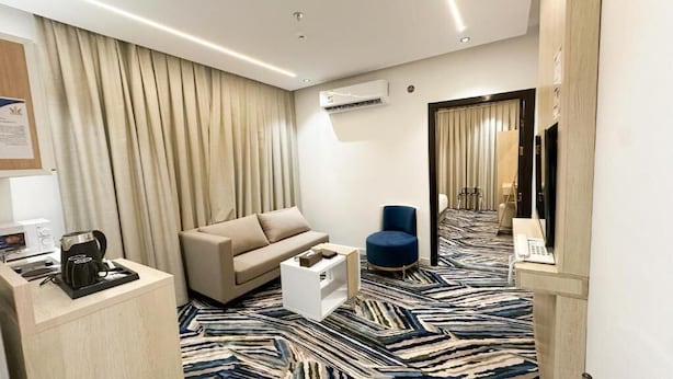 Une chambre d’hôtel avec un canapé beige, un fauteuil bleu, une machine à café et un tapis à motifs.