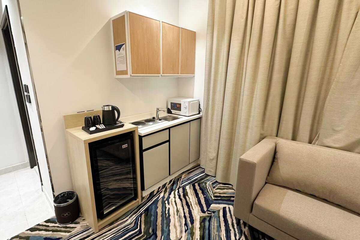 A compact hotel room with a kitchenette, a sofa, and a patterned rug.