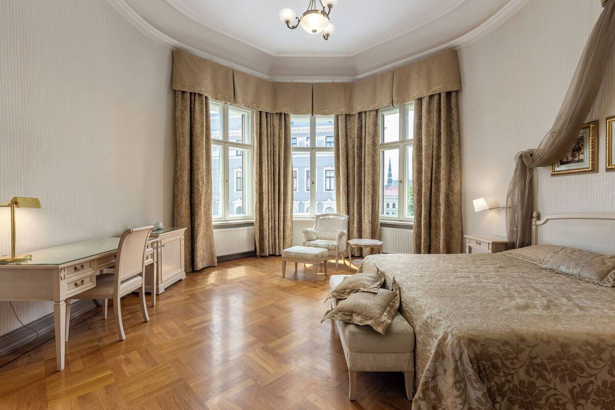 A hotel room with a large bed, a desk, a chair, and a small table. There are two windows with curtains.