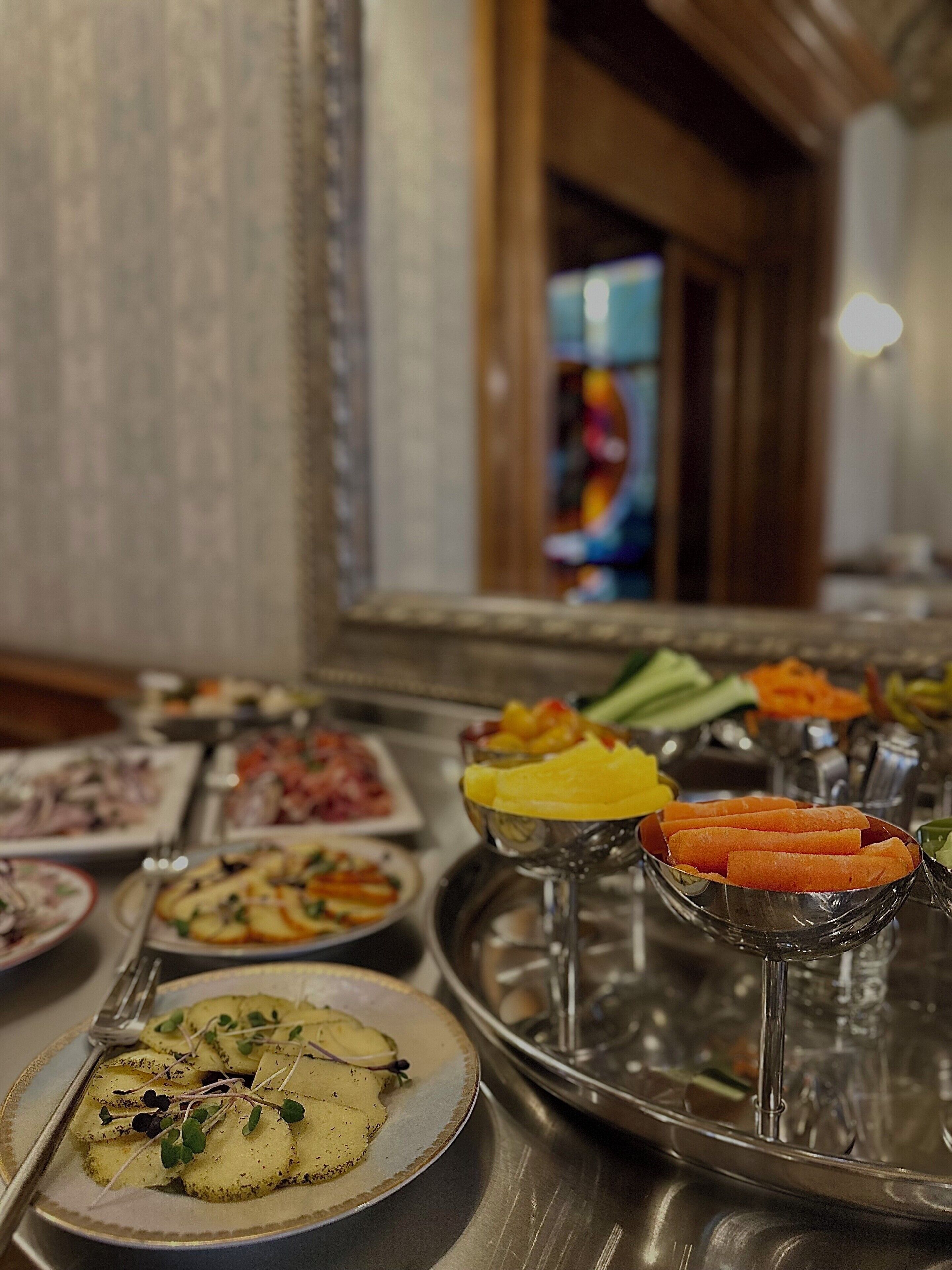 A buffet table with various dishes, including a plate of sliced potatoes garnished with greens, a bowl of sliced carrots, and a tray with fruits and vegetables.