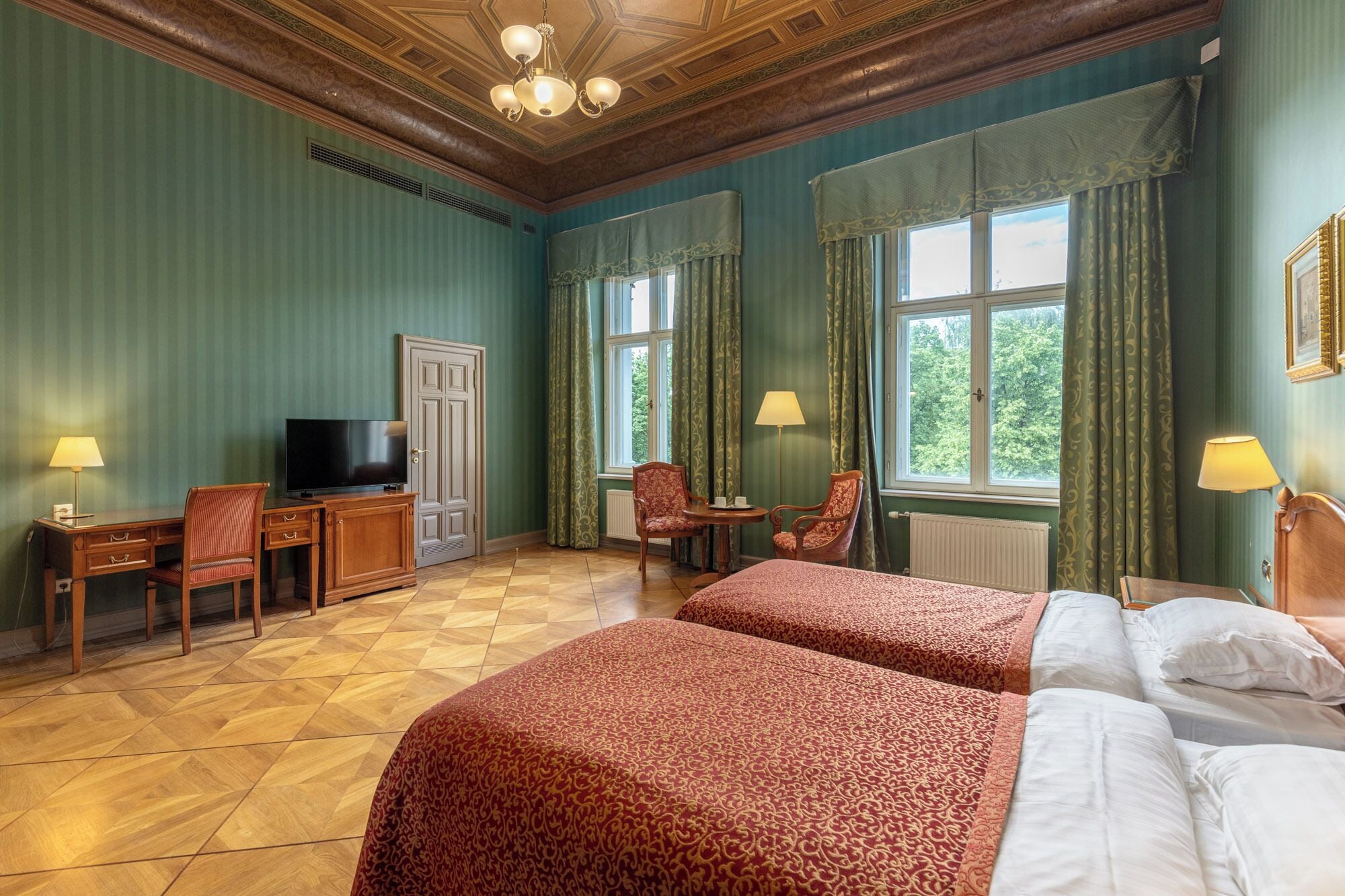 Quarto de hotel com duas camas, uma secretária com televisão, duas cadeiras e janelas amplas com cortinas.