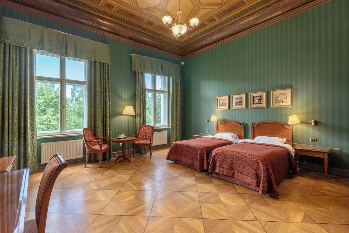 A hotel room with two beds, a sitting area with chairs, a table, and a view of trees through the windows.
