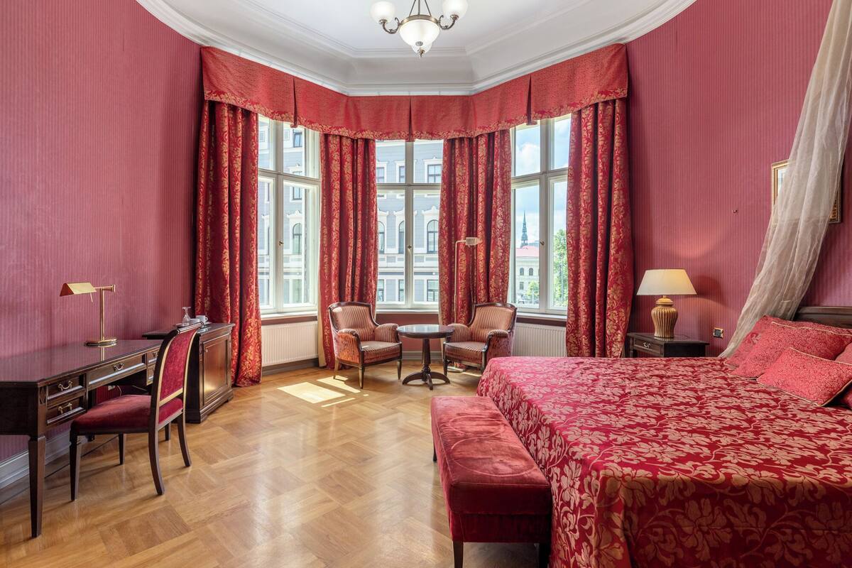 A hotel room with a large bed, two armchairs, a desk with a lamp, and a view of buildings through large windows.
