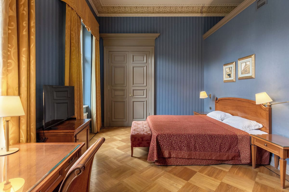 A hotel room with a wooden bed, a desk with a lamp, a television, and a wardrobe.