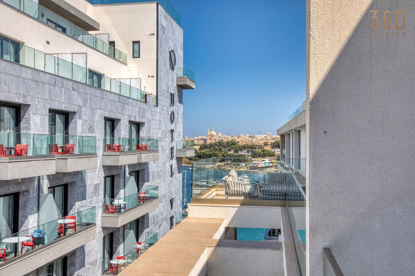 Un hotel moderno con varios balcones, mobiliario exterior y vistas a un paisaje urbano histórico.