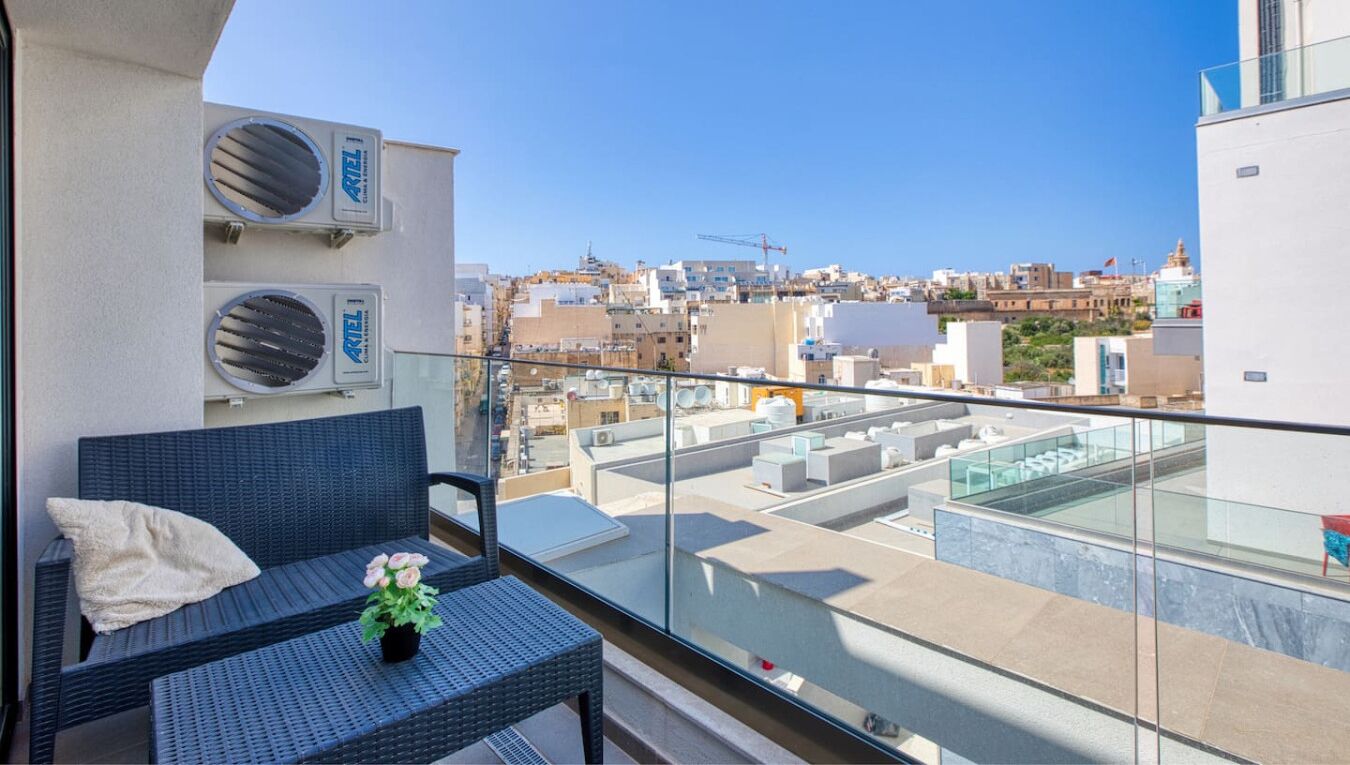 Un balcon avec un fauteuil en osier, une petite table ornée d’une fleur et une vue sur le paysage urbain, avec ses bâtiments et ses grues de construction.