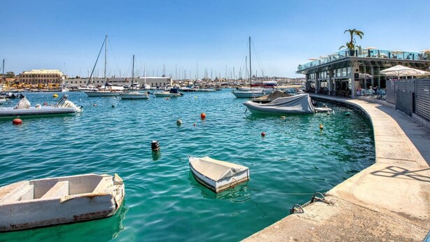 Un puerto deportivo con barcos amarrados, un restaurante frente al agua y vistas de la ciudad al fondo.