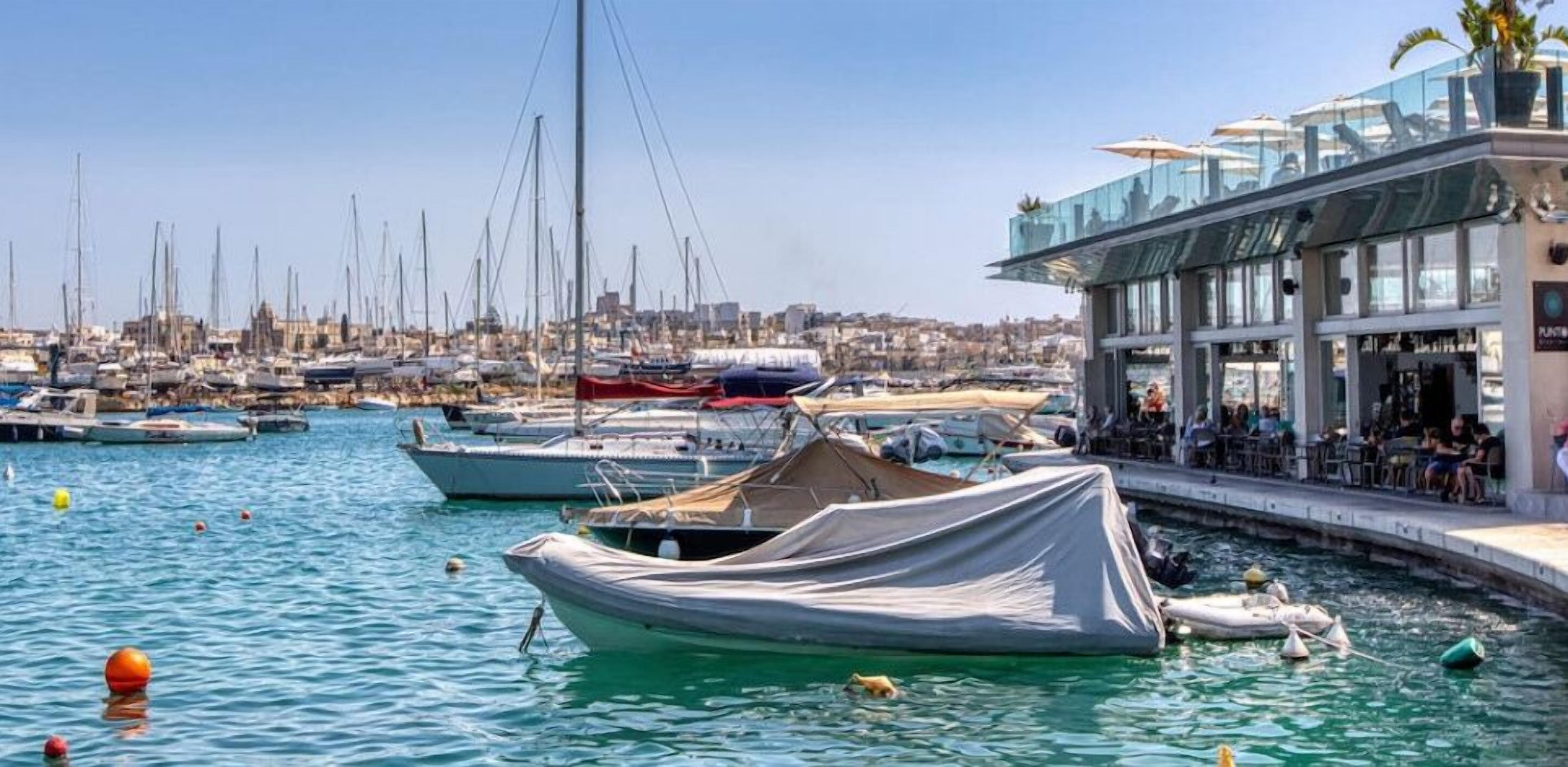 Marina con barcos amarrados, un edificio con techo de cristal y un paisaje urbano al fondo.