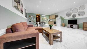 A modern living room with a sofa, a coffee table, and a TV. There is a kitchen in the background with cabinets and a refrigerator. - An amazing 3BR maisonette with private terrace &AC (Marsaskala)
