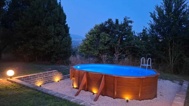 Una piscina elevada de madera con escalera, rodeada por un camino de grava e iluminada por luces de jardín.
