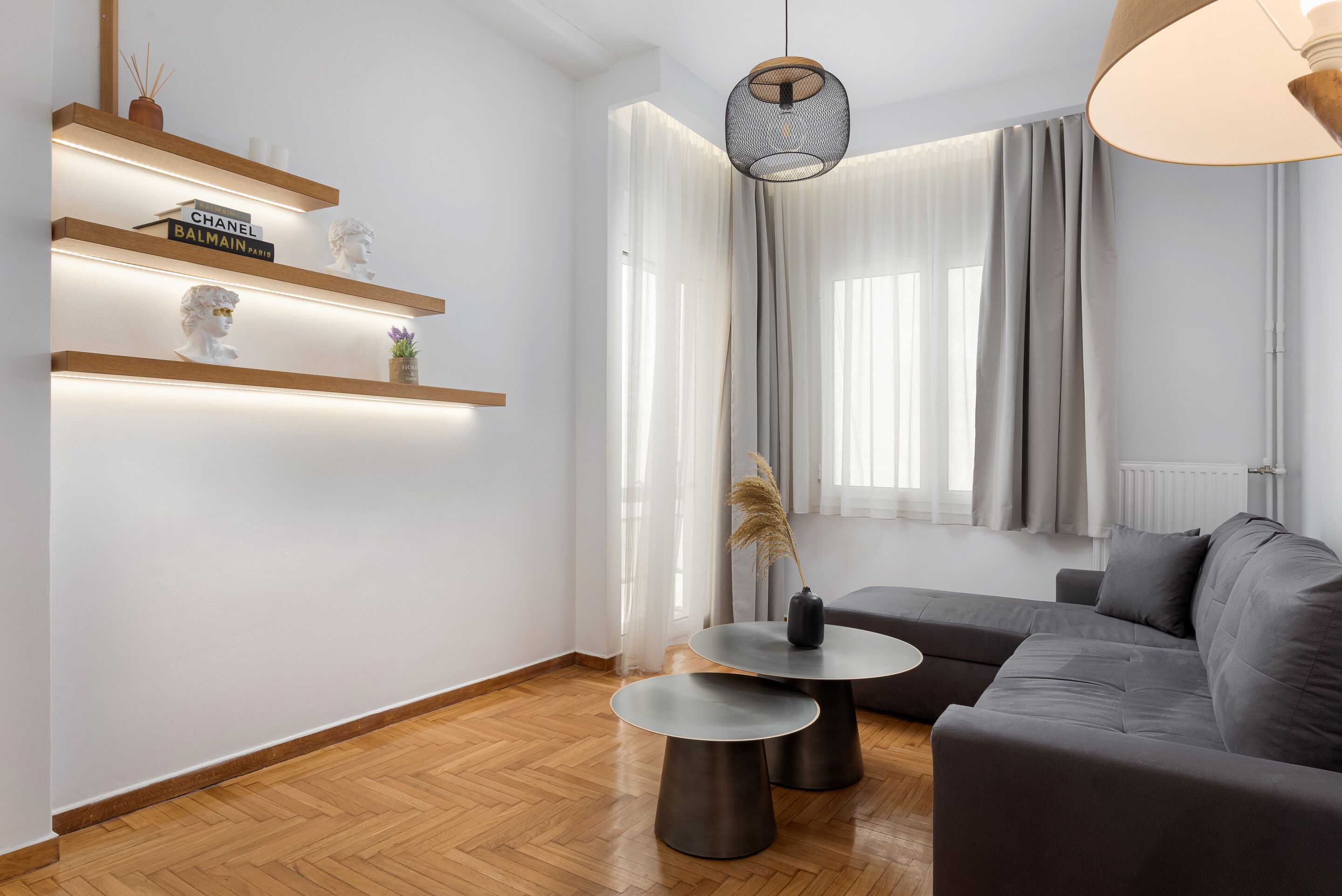 A modern living room with a grey sofa, two round tables, and a wooden floor. There are shelves with decorative items and a hanging light fixture.