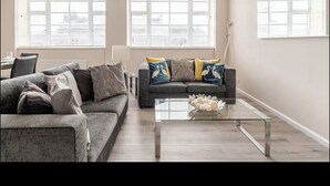 Un salon moderne avec un canapé gris, une table basse en verre et une grande fenêtre équipée de stores.