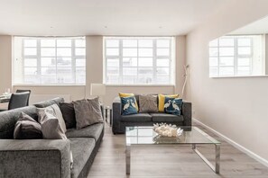 A modern living room with a grey sofa, a glass coffee table, and a large window with blinds. - Brighton central perfect long stays with free parking. (Brighton)