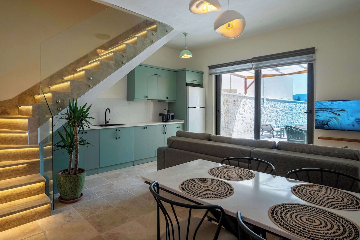 A modern kitchen with teal cabinets, a dining area with a table and chairs, and a staircase with illuminated steps.