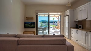 En moderne stue med en sofa, et fjernsyn og en skydedør, der fører ud til en terrasse med havemøbler.