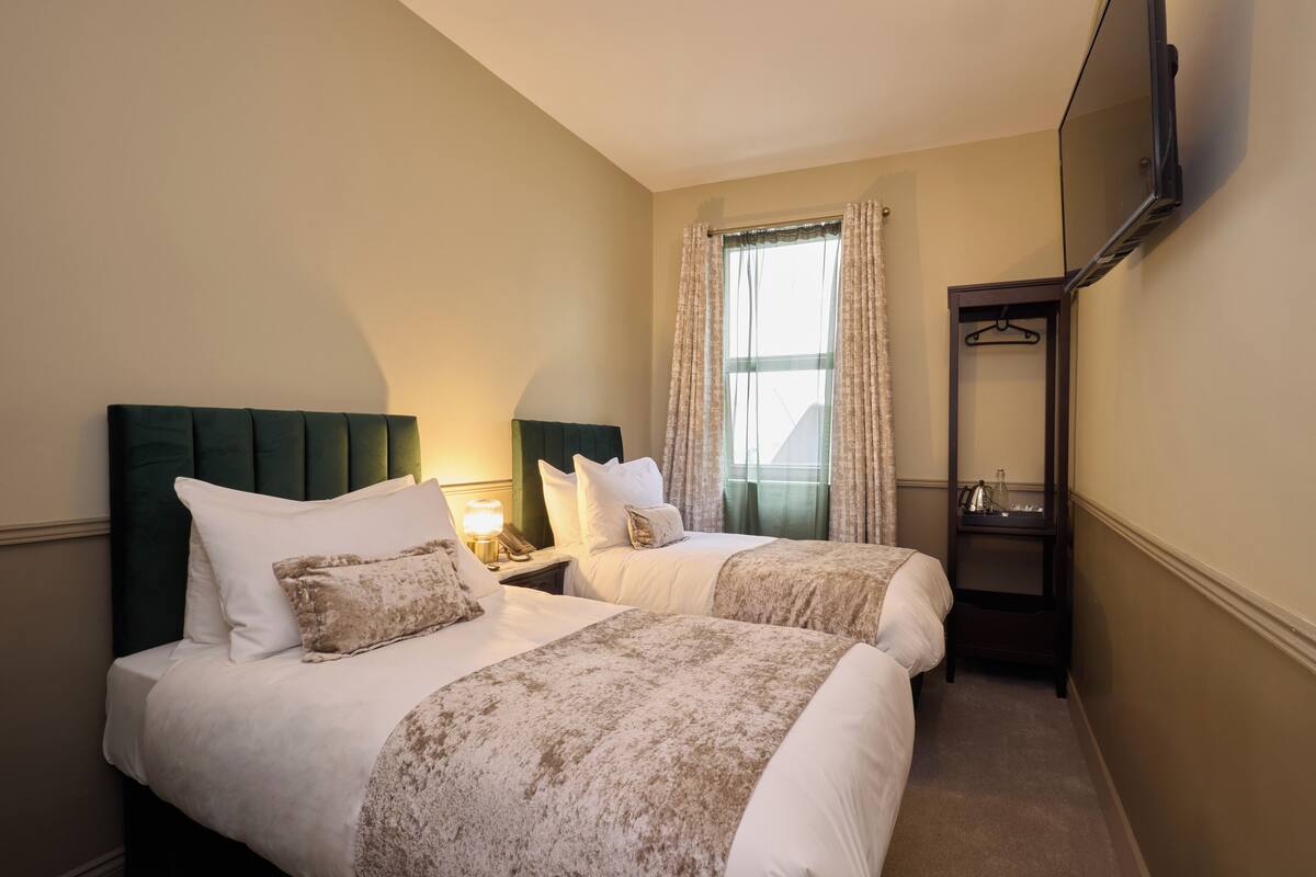 A hotel room with two beds, a TV mounted on the wall, a window with curtains, and a bedside table with a lamp.