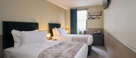 A hotel room with two beds, a TV mounted on the wall, a bedside table with a lamp, and a window with curtains.