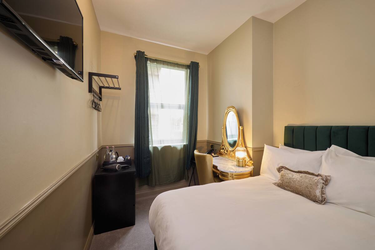 A hotel room with a bed, a desk with a mirror, a chair, a small table with toiletries, and a window with curtains.