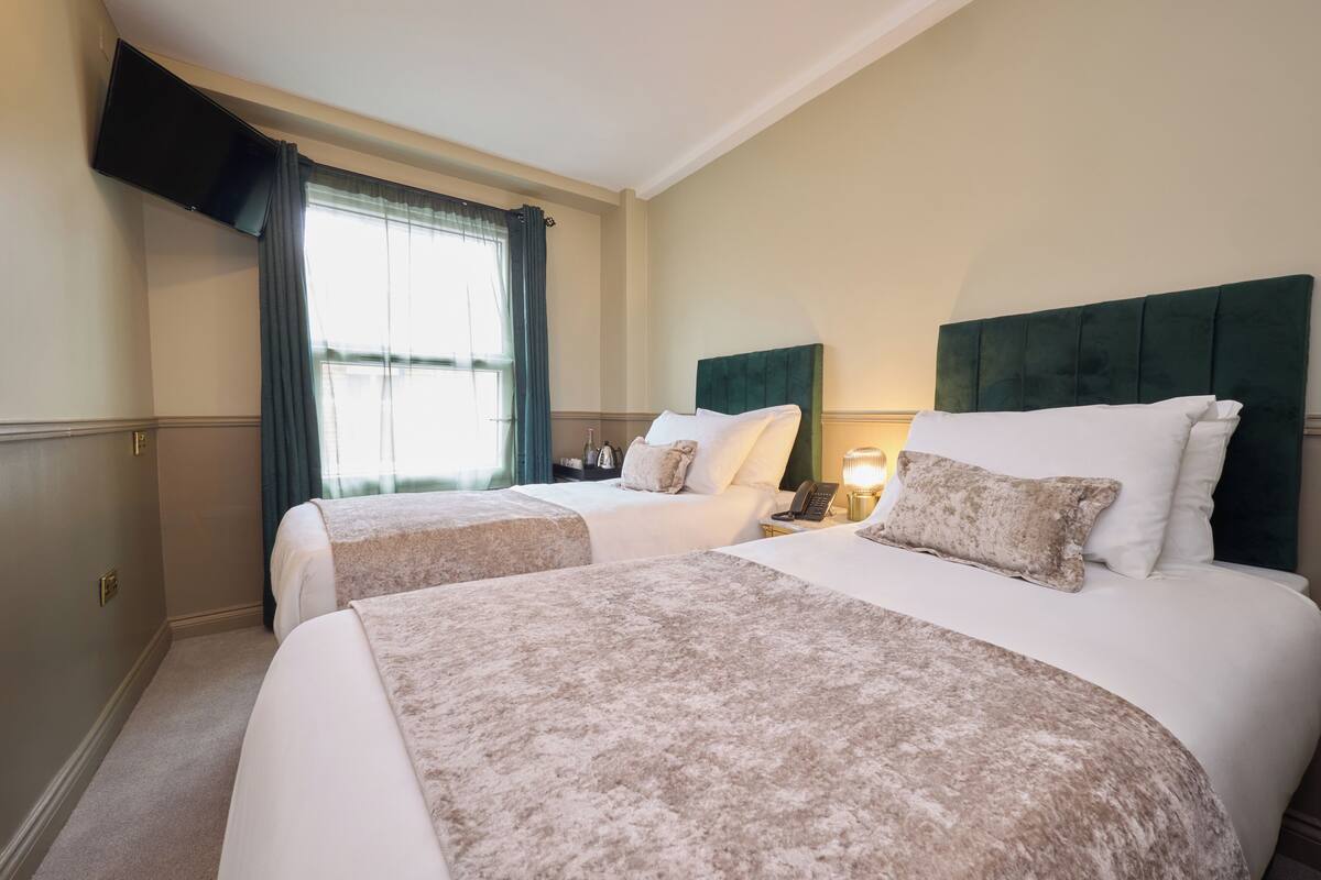 A hotel room with two beds, a flat-screen TV mounted on the wall, a window with curtains, and a bedside table with a lamp.