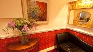 Una habitación con pared de color rojo con estampado, un sillón de cuero negro, una mesita de madera con un jarrón de flores y un espejo grande con marco dorado.