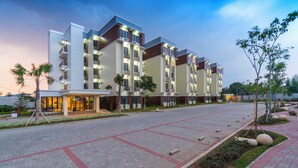Un hôtel moderne à plusieurs étages, doté d’un espace extérieur bien entretenu avec des allées pavées et des palmiers.