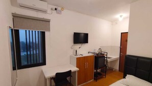 Une chambre d’hôtel de taille réduite, comprenant un lit, un bureau avec une chaise, une télévision, une armoire et une fenêtre avec des stores.