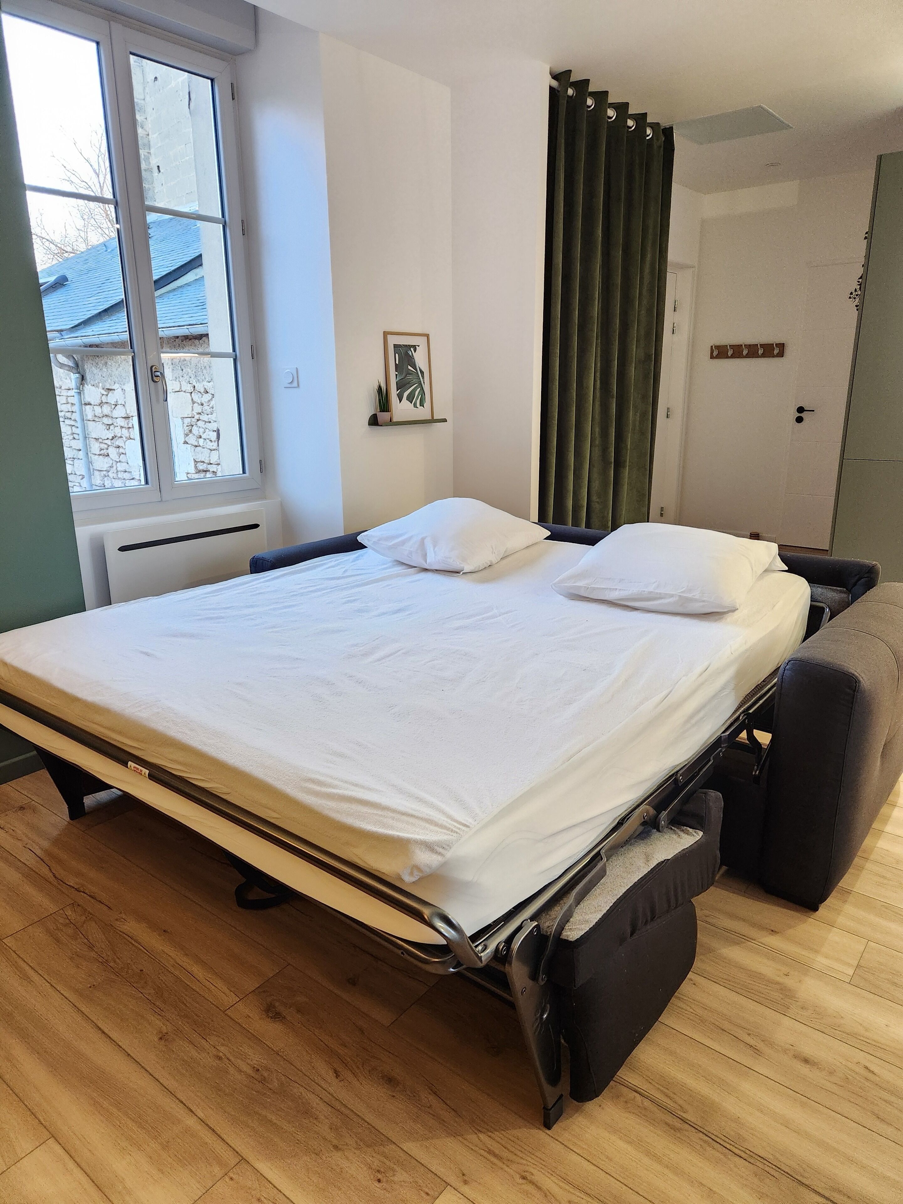 Een netjes opgemaakt bed met een witte matras en kussens, een raam met gordijnen, een radiator en een klein fotokader aan de muur.