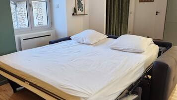 Een netjes opgemaakt bed met een witte matras en kussens, een raam met gordijnen, een radiator en een klein fotokader aan de muur.