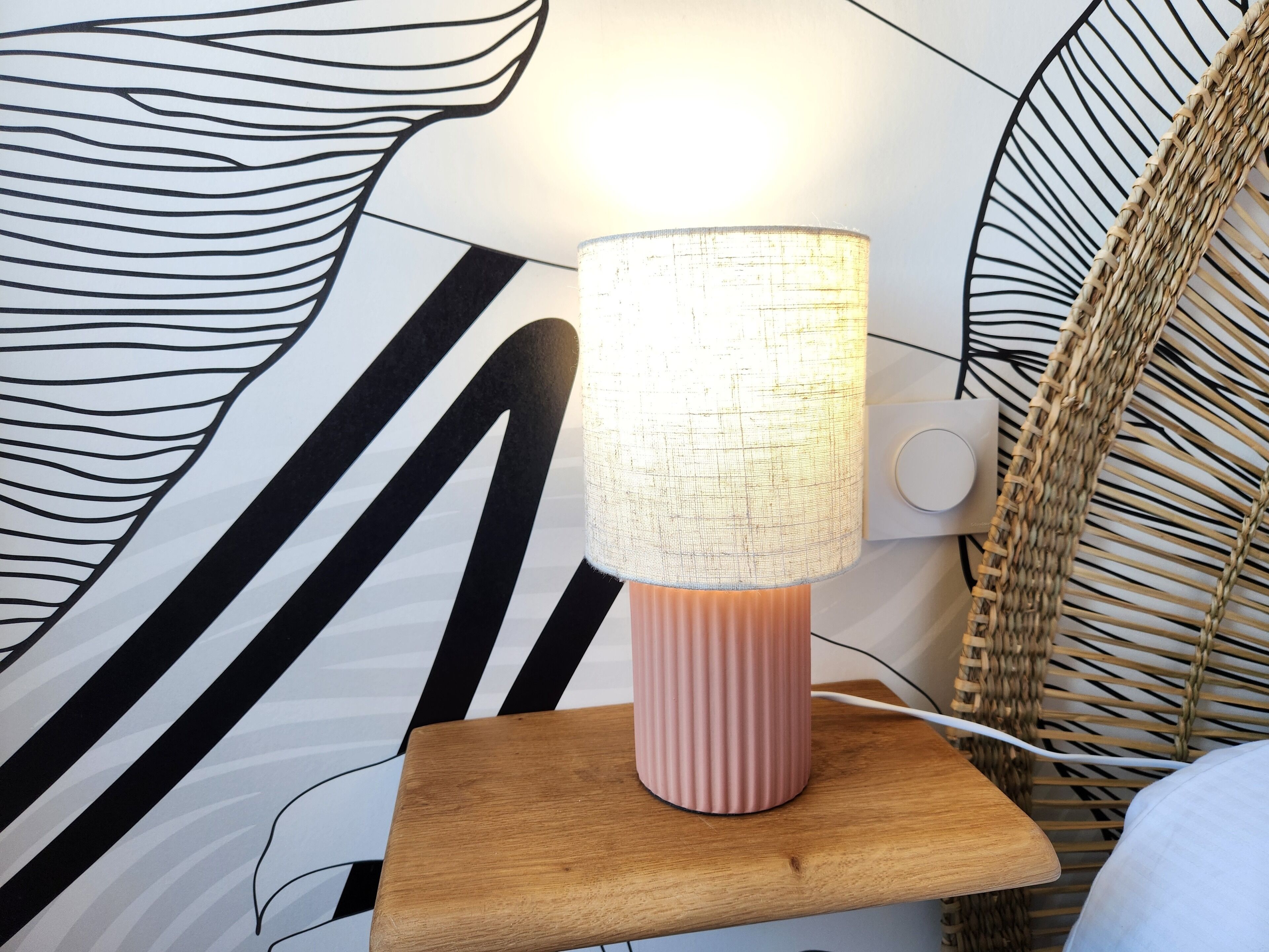A lamp on a wooden nightstand with a textured lampshade, set against a wall with a black and white abstract design.