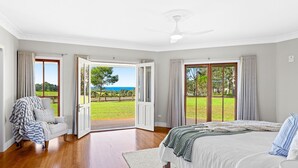 A bedroom with a large bed, a view of a grassy area through a sliding door, and a ceiling fan.