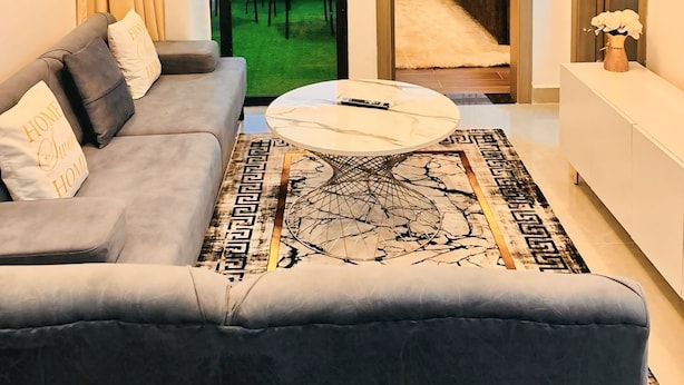 A modern living room with a grey sofa, a round coffee table, and a view of a cityscape through a glass door. - Valley Heights Apartments (Nairobi)