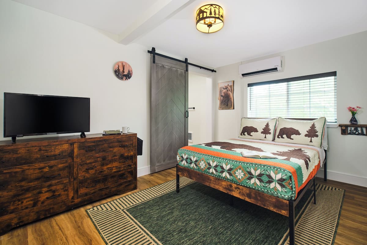 Un dormitorio con un aparador de madera, una cama con colcha, un televisor sobre el aparador, una ventana con persianas y un reloj de pared.