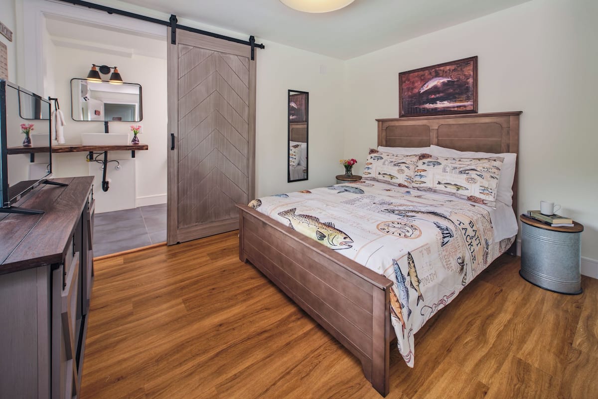 Un dormitorio con una cama de madera, una mesita de noche con una lámpara, un espejo, una puerta corredera y una pequeña mesa redonda con una taza y un libro.